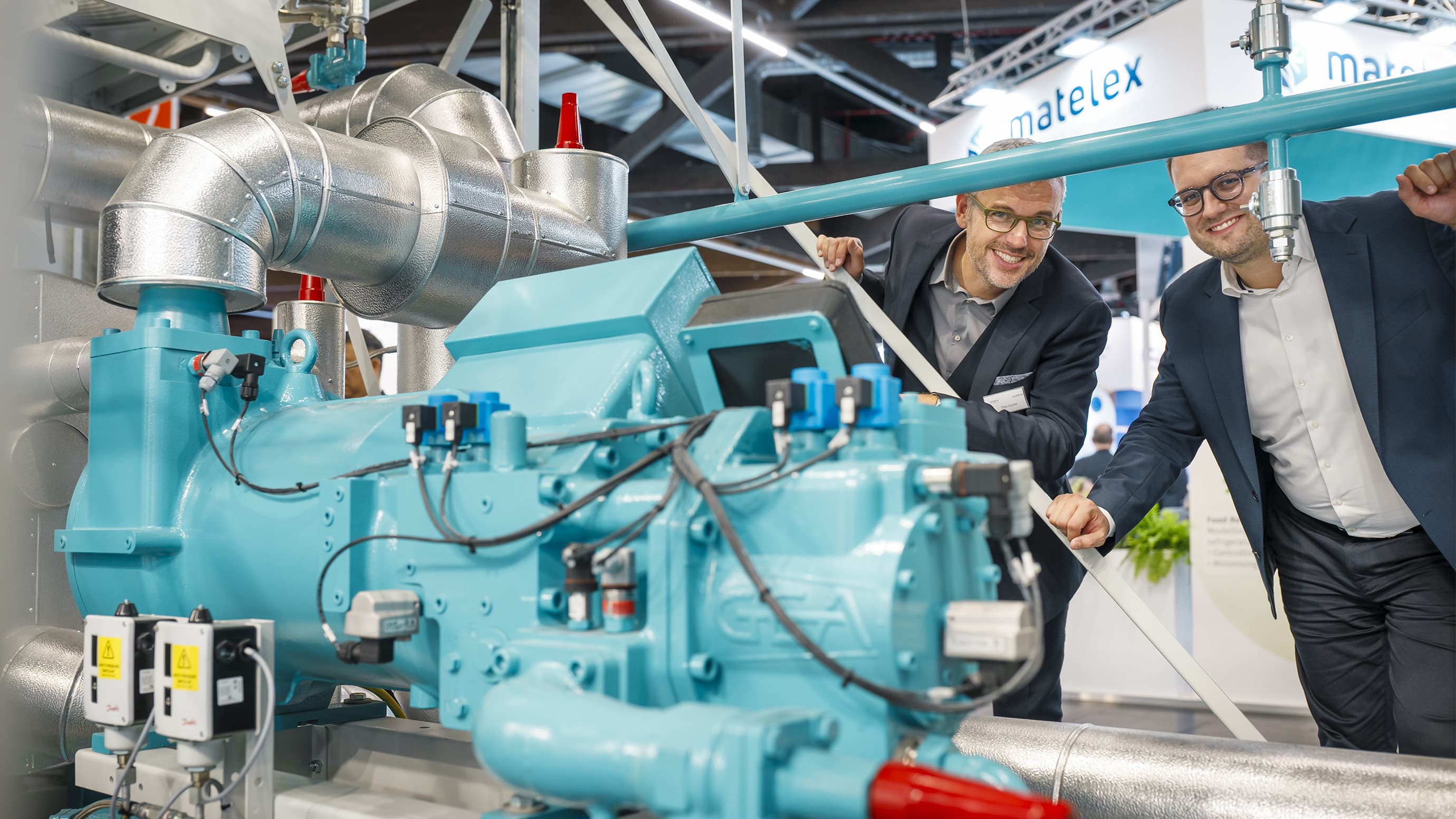 Two male exhibition participants smiling in the camera while standing behind a refrigeration machine